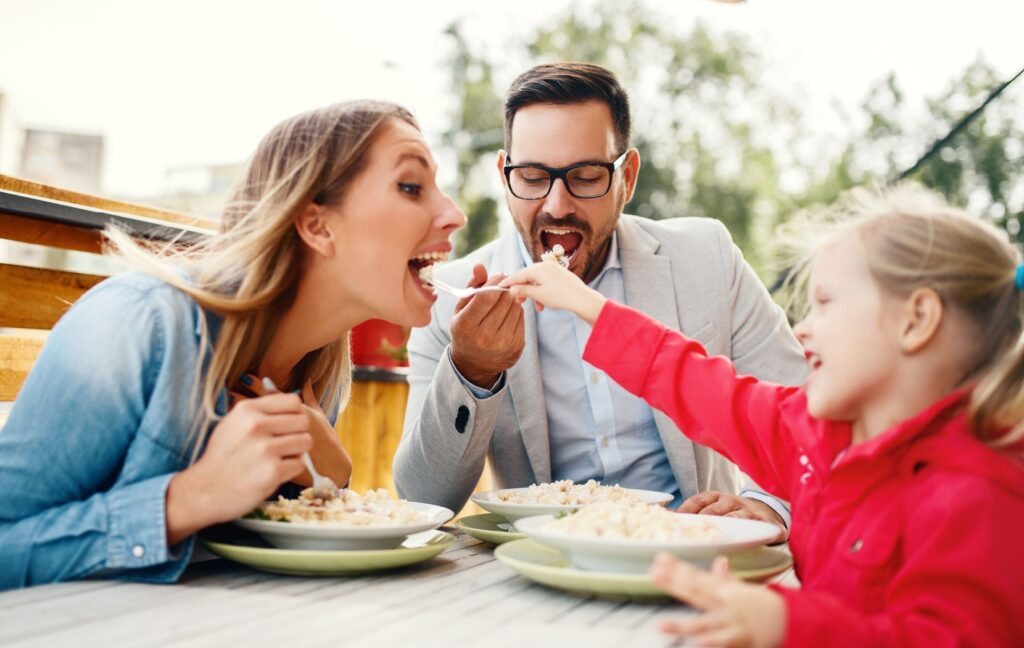 A parent eats food from their smiling child's fork, increasing the risk of transmitting cavity-causing bacteria.