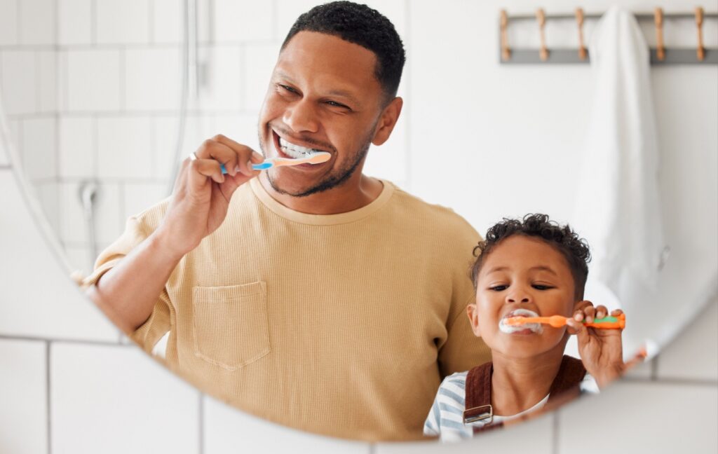 A parent and a small child stand side by side in front of a bathroom mirror while they carefully brush their teeth.