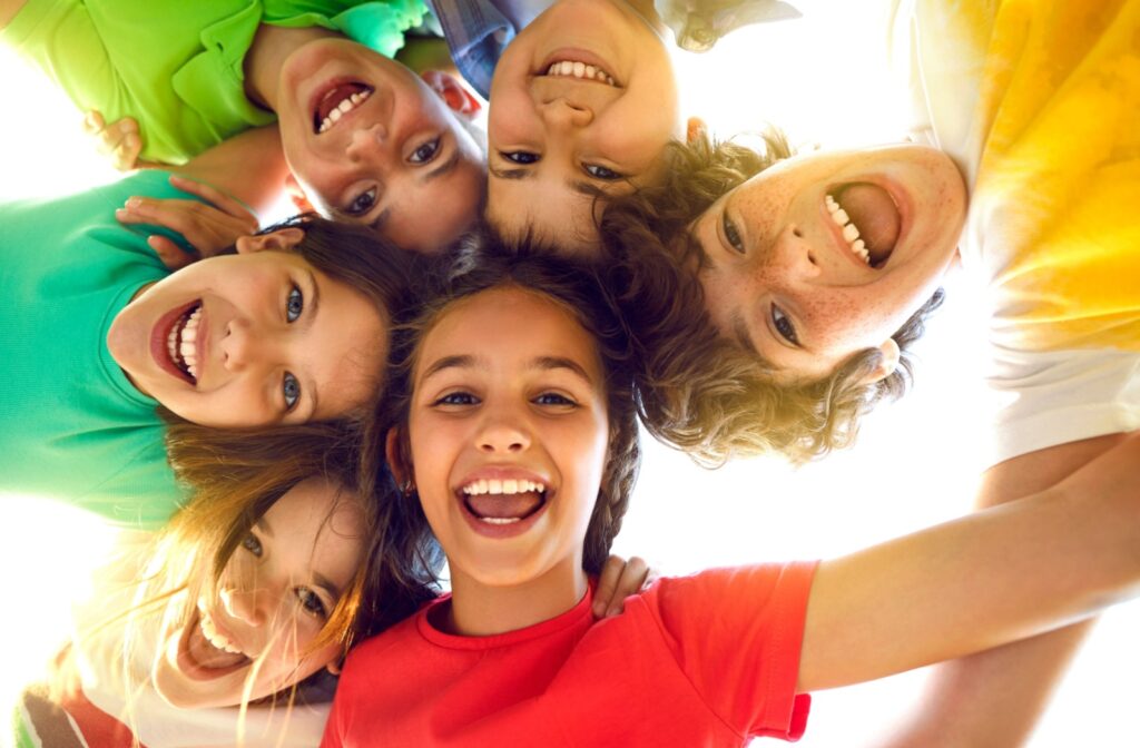 A group of six joyful children with bright smiles form a circle looking down at the camera. They're wearing colorful t-shirts in green, red, white, and yellow, creating a cheerful huddle. The children's faces show diverse features and their expressions convey happiness and friendship as sunlight filters through from above, giving the image a warm, positive atmosphere.