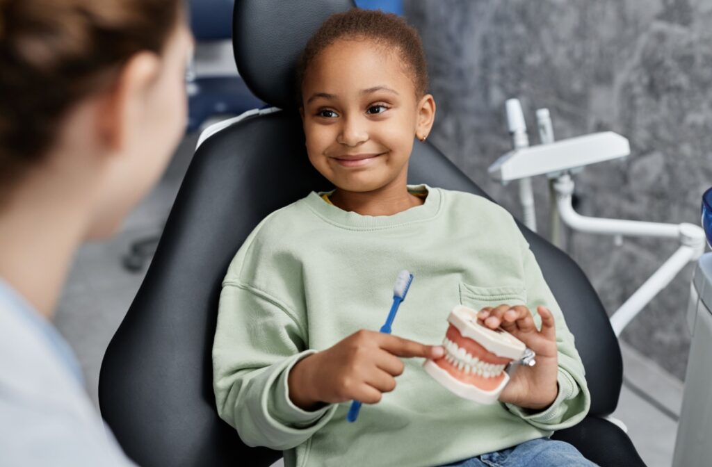 A young child with a warm smile sits in a dental chair while holding a dental model and a toothbrush. The child is wearing a light mint green sweatshirt and appears to be learning about proper dental care during a pediatric dental appointment. A dental professional can be partially seen in the foreground.