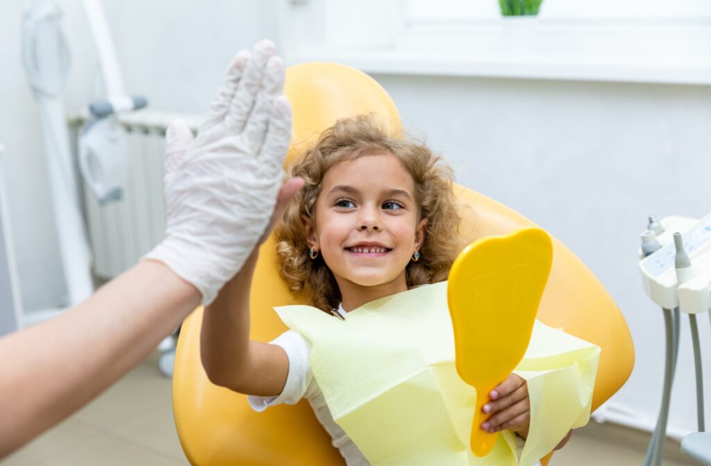 Smiling young girl with curly hair sitting comfortably in a bright yellow pediatric dental chair at London Little Bites Dentistry, with a dentist's gloved hand visible and dental equipment in the background.