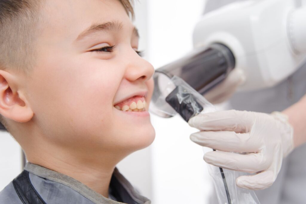 Young boy smiling during dental examination while dentist takes bitewing x-ray of teeth.