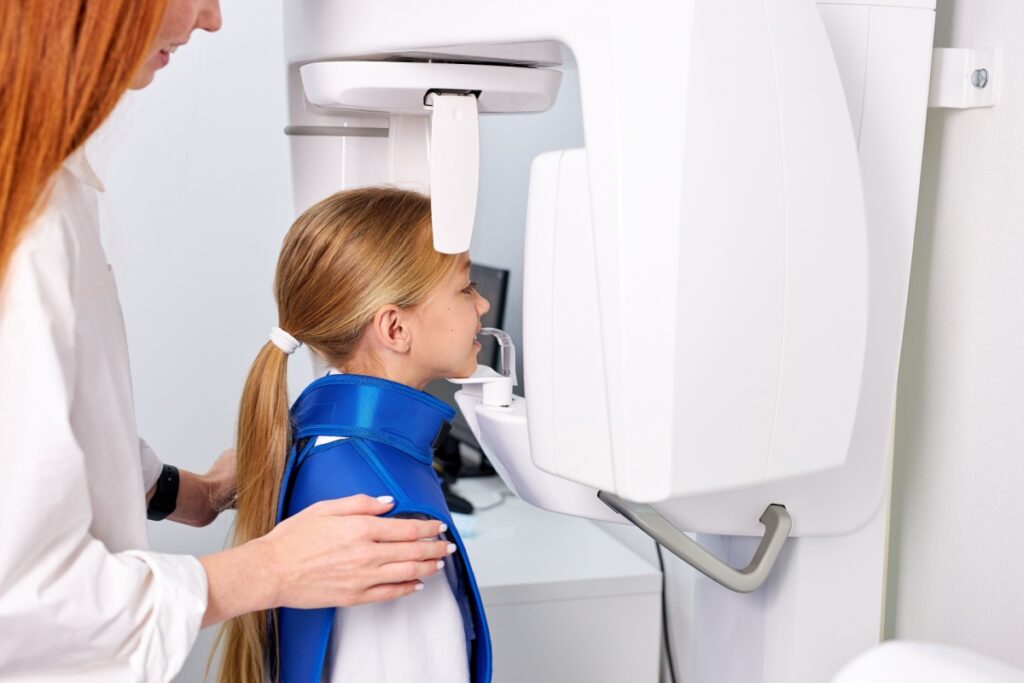 Young girl positioned for panoramic dental x-ray imaging with dental professional assistance.