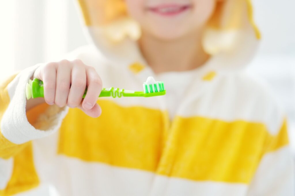 Young child in yellow and white clothing holding green toothbrush demonstrating proper oral hygiene habits.
