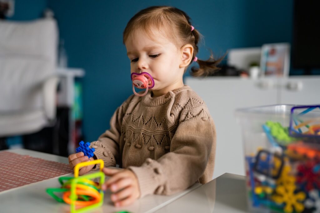 Toddler girl using pacifier and playing with toys.