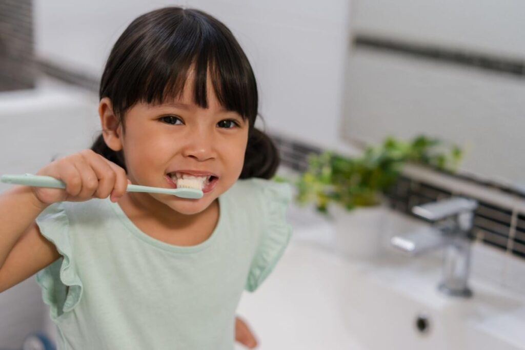 A young Asian girl with a bright smile brushing her teeth with a light green toothbrush in a clean bathroom setting at London Little Bites Dentistry.